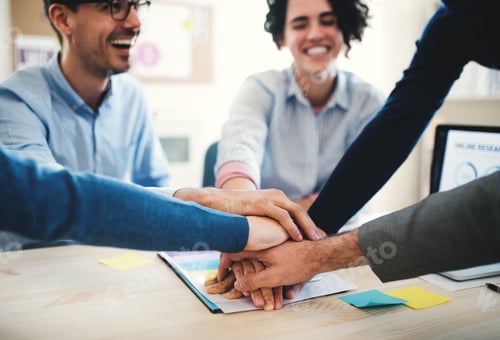 Preview: Young businesspeople sitting around table in a modern office, stacking hands.