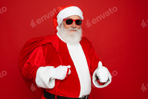 Preview: Santa Claus in sun eyeglasses with sack bag demonstrate thumb up isolated red color background