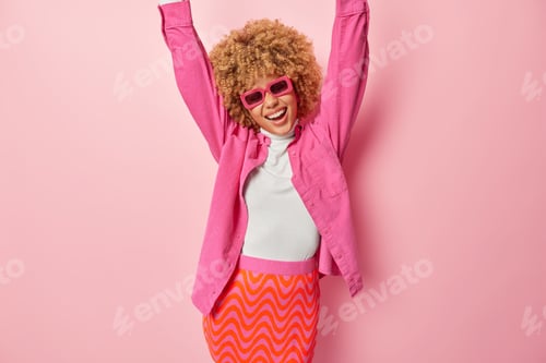 Preview: Horizontal shot of pretty joyful young woman with curly hair keeps arms raised up laughs happily has