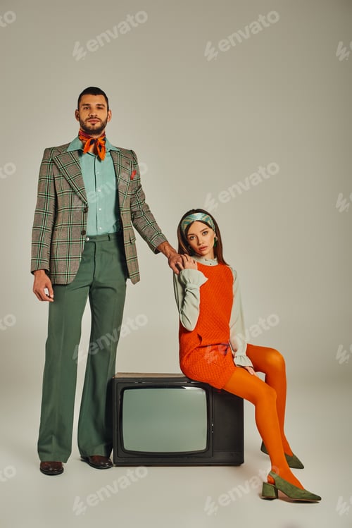 Preview: confident man holding hands with charming woman sitting on vintage tv set on grey, retro lifestyle