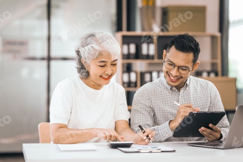 Preview: An older mature woman shows something to asian man on laptop. smile and work together in office.