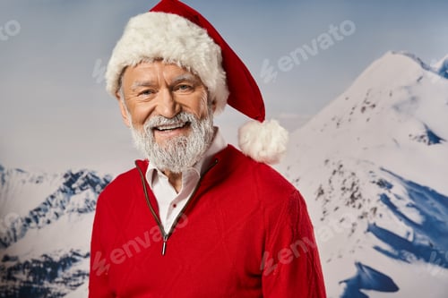 Preview: good looking jolly Santa in red attire smiling at camera with mountain backdrop, winter concept