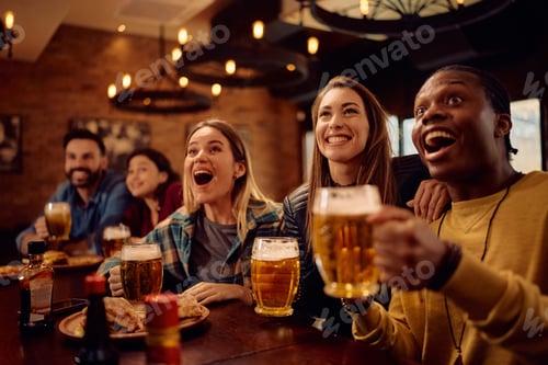 Preview: Multiracial group of friends drinking beer and watching sports game on TV in a bar.