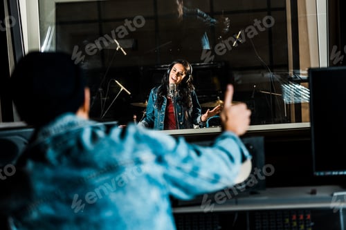 Preview: selective focus of mixed race sound producer showing thumb up to woman singing in recording studio