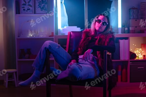 Preview: Beautiful girl looking to camera with sun glasses and fashion outfit in a room with colorful neon