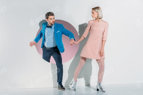 Preview: young couple in suit and dress holding hands and looking at each other on grey
