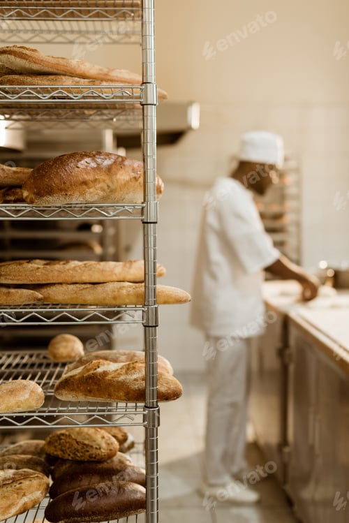 Preview: african american baker preparing raw dough at workplace with shelves of fresh bread on foreground