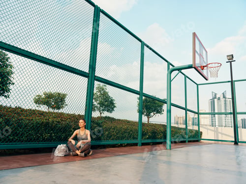 Preview: Young woman resting on a basketball court under a clear blue sky, with a hoop in the background and