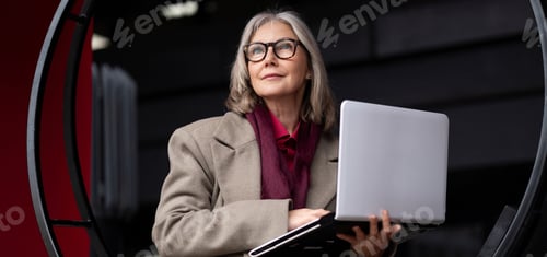 Preview: Elderly businesswoman stands outside her office while holding a laptop during daylight hours