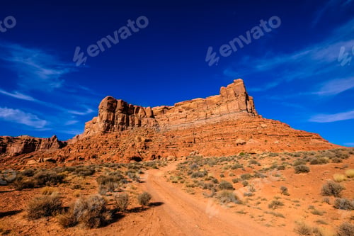 Preview: Dirt road in the middle of the desert with dried bushes and a cliff in the distance under blue sky