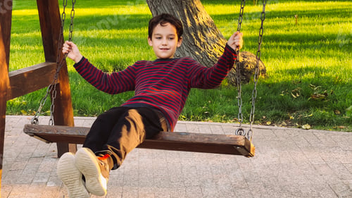 Preview: A young boy with brown hair sits on a wooden swing in a park.