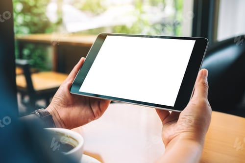Preview: Hands holding black tablet pc with blank white screen with coffee cup on wooden table