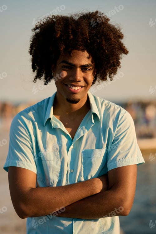 Preview: Young man smiling with folded arms on beach at sunset