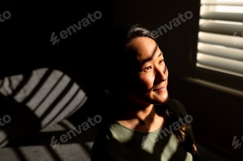 Preview: Portrait of happy asian woman standing in ray of light at window