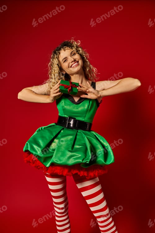 Preview: joyful curly woman dressed as new year elf smiling sincerely and showing present at camera
