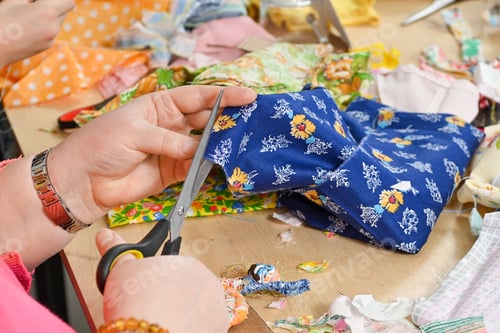 Preview: Womans Hands Cutting Blue Fabric with Silver Scissors on Table with Fabric Scraps in Sewing Studio
