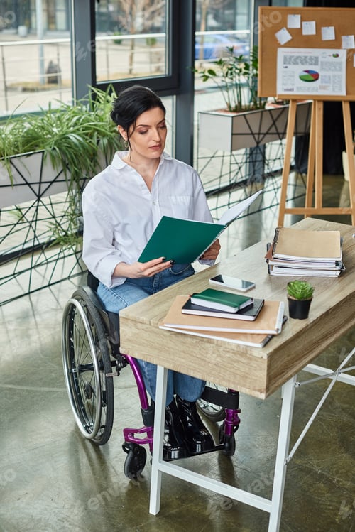 Preview: dedicated businesswoman with mobility disability in wheelchair looking at her paperwork in office