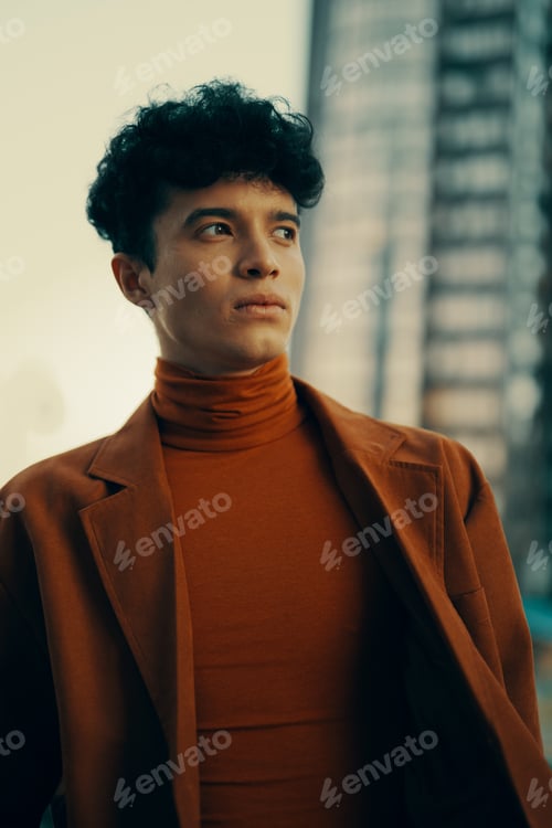 Preview: Thoughtful young man in a stylish rust colored outfit against a blurred cityscape backdrop