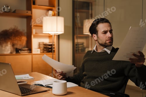 Preview: Young puzzled man working with laptop and papers