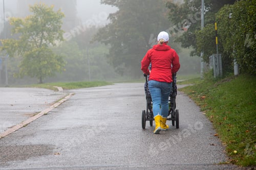 Preview: Person wearing jeans paired with yellow boots and a red coat walking with a baby carriage in a park