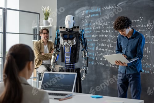 Preview: Clever teenage guy with laptop standing by blackboard in front of class