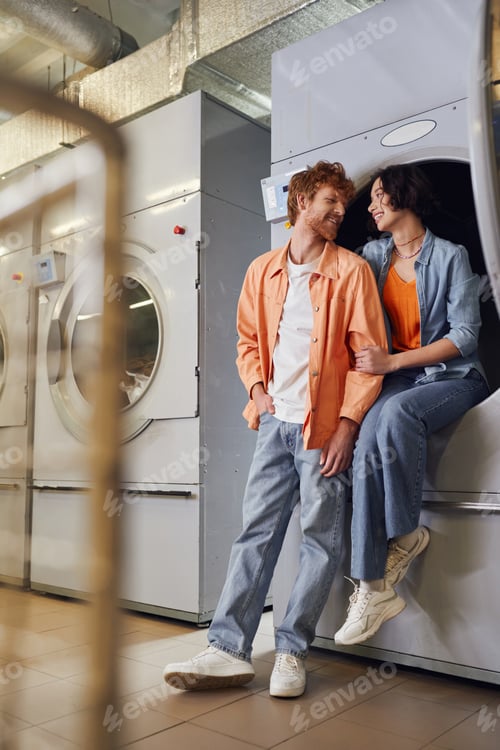 Preview: full length of cheerful multiethnic couple talking near washing machine in public laundry