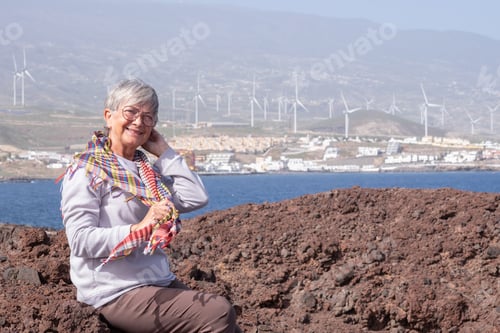Preview: Portrait of carefree senior woman sitting outdoor at sea wearing eyeglasses looking at camera