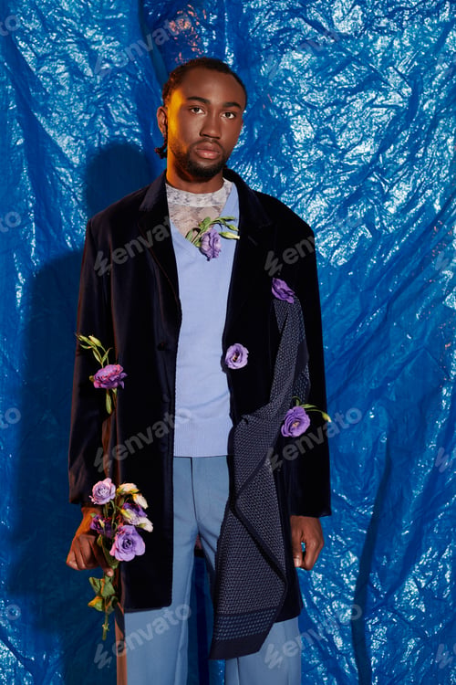Preview: Handsome young man adorned with flowers poses confidently against a vibrant blue backdrop