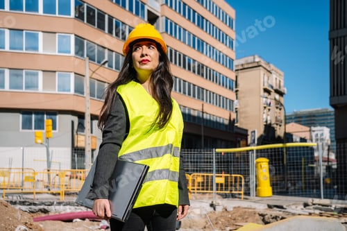 Preview: Ethnic female inspector in safety vest standing near building with laptop in hand
