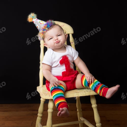 Preview: Little girl makes a funny face while sitting on a yellow chair. Birthday hat, colorful striped socks