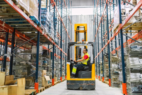 Preview: Warehouse worker driving forklift between tall shelves full of goods