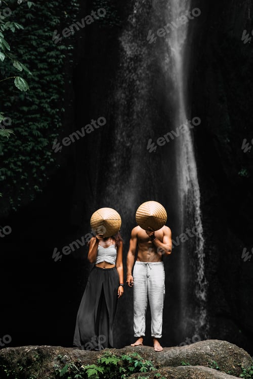 Preview: A couple stands at a waterfall and covers their faces with rice caps