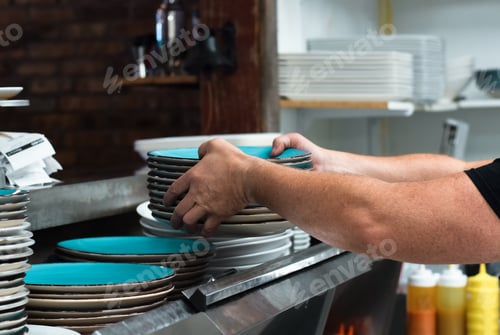Preview: Restaurant kitchen employee placing stack of clean dishes on a shelf