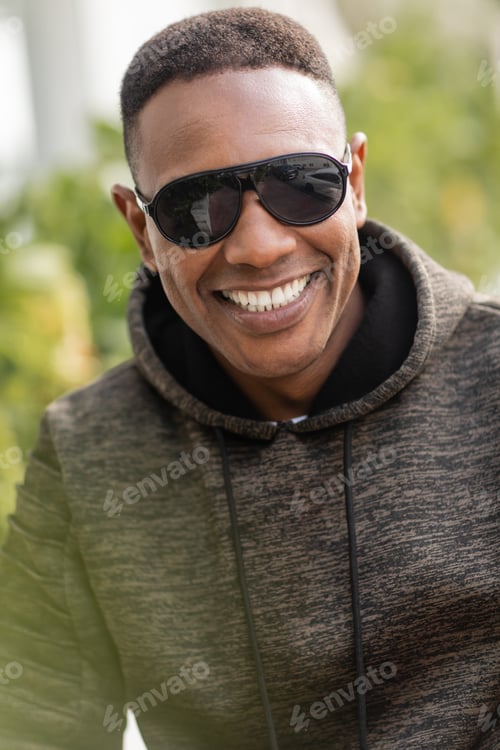 Preview: portrait of positive african american man in hoodie and stylish sunglasses smiling outdoors