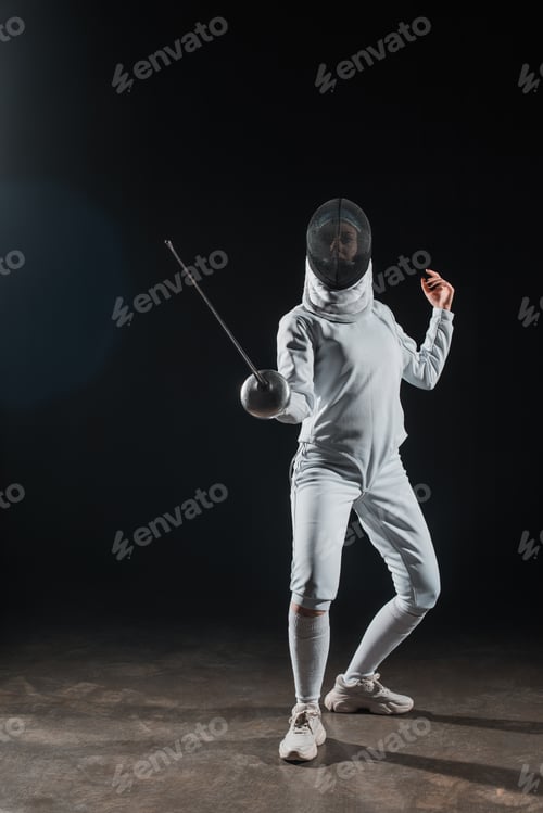 Preview: Fencer in fencing mask holding rapier while training under spotlight on black background