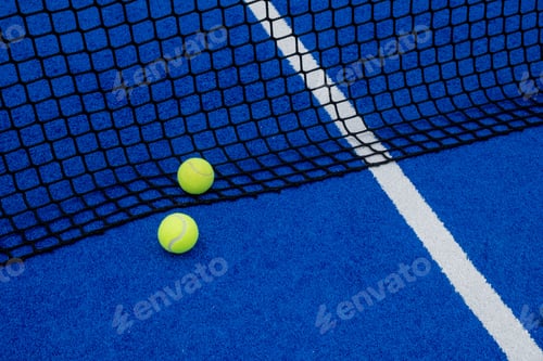 Preview: View of two balls near the net of a blue artificial grass paddle tennis court.
