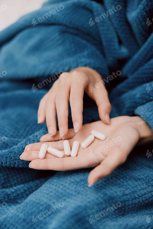 Preview: Woman sitting on bed wrapped in blue blanket, holding pills in hands