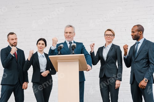 Preview: happy speaker in glasses and multicultural businessmen and businesswomen showing clenched fists