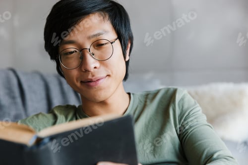 Vista previa: Image of young asian man wearing eyeglasses reading book in apartment