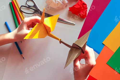 Preview: Children’s hands doing origami crane from yellow paper on white background