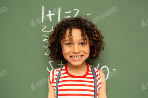 Preview: Front view of a cute mixed-race schoolgirl standing against green board in a classroom at school