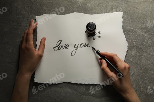 Preview: Ink bottle, pen in hand and words Love you on paper on gray background, top view