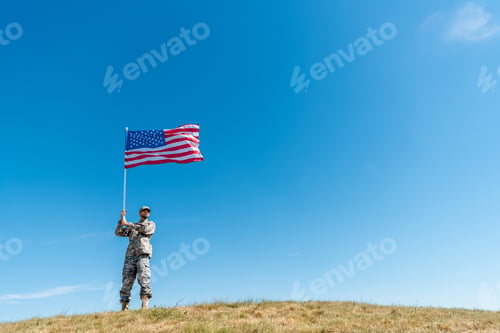 Preview: low angle view of handsome military man in uniform walking with american flag with stars and stripes