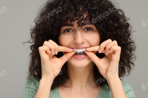 Preview: Young woman applying whitening strip on her teeth against grey background