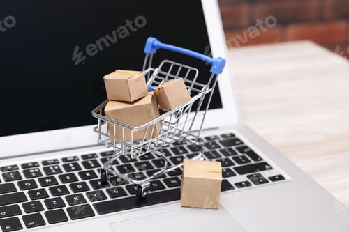 Preview: Internet store. Small cardboard boxes, shopping cart and laptop on light wooden table, closeup