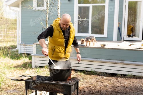 Preview: A man in a yellow tank top cooking on a fire in a cauldron in the backyard of the house