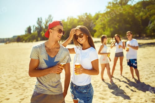 Preview: Smiling young couple clinking glasses with beer while people barbecuing in the background.