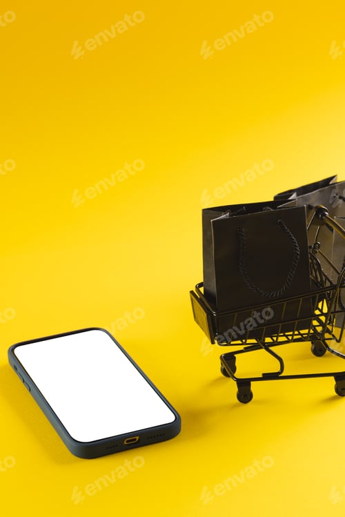 Preview: Vertical image of smartphone with copy space and trolley with bags on yellow background