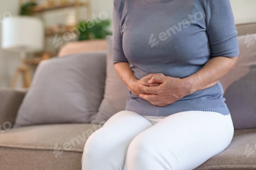 Preview: Old woman suffering stomach ache sitting on couch in living room at home.