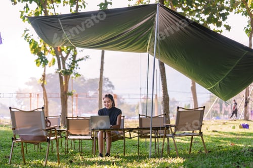 Preview: Young asian freelancer woman is working with laptop under tarp in park during sunny summer morning,